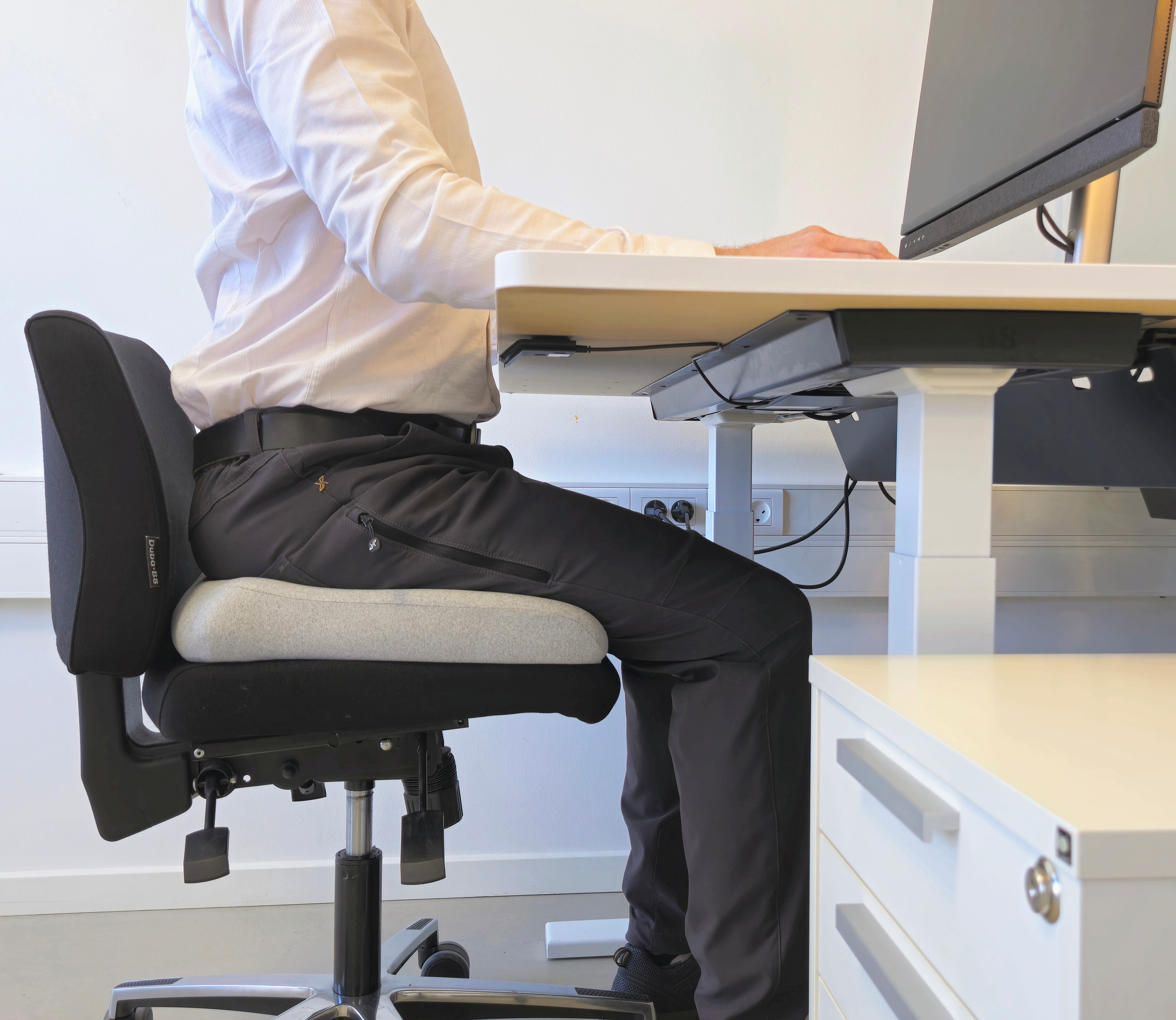 Mand sidder ved skrivebord på ergonomisk grå memory foam siddepude fra Copenhagen Comfort, giver støtte til ryg og hofter under kontorarbejde.